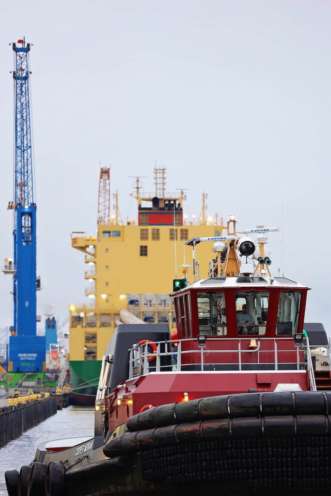 A red tugboat chugs directly towards the camera in the foreground while a yellow ship with a green hull is docked in the background next to a bright blue crane