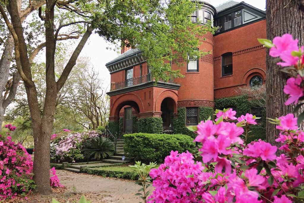 Stately 3-story brick mansion in the background with pink, hot pink, and white azaleas near the front entrance, and dozens of hot pink blooms in the foreground of the photo