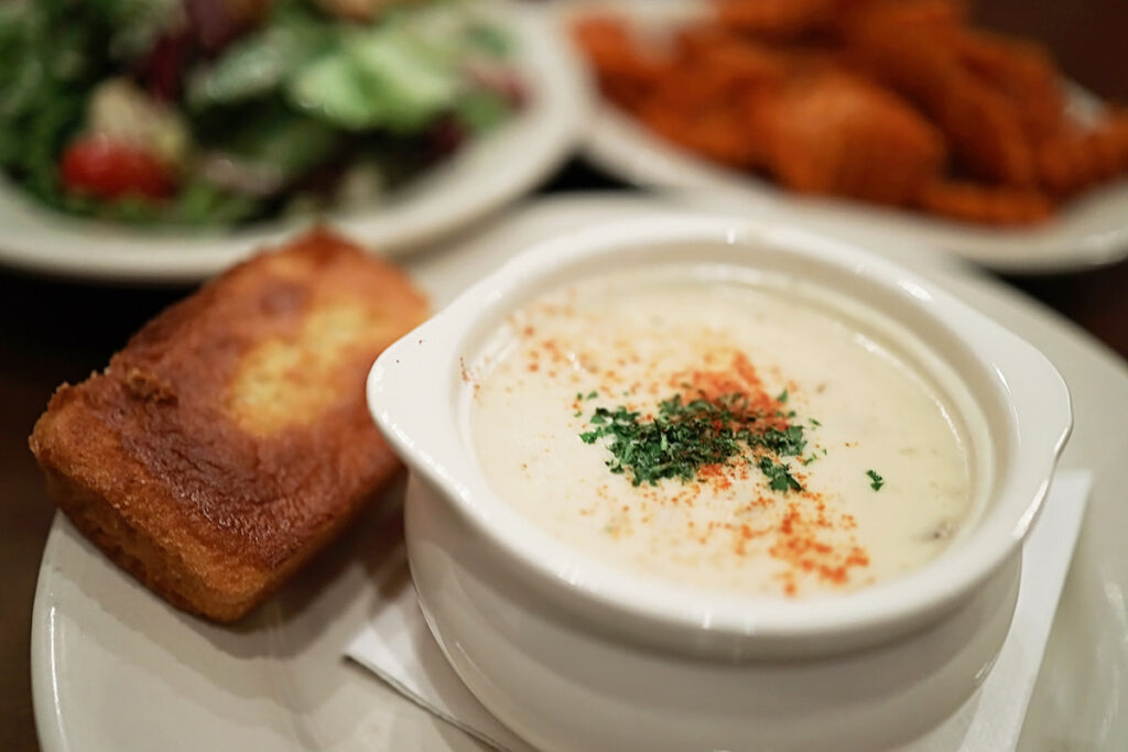 White cup filled with creamy crab soup. Orange and green seasonings are sprinkled across the top. A golden mini-loaf of cornbread, a salad, and a plate of sweet potato fries are visible in the background