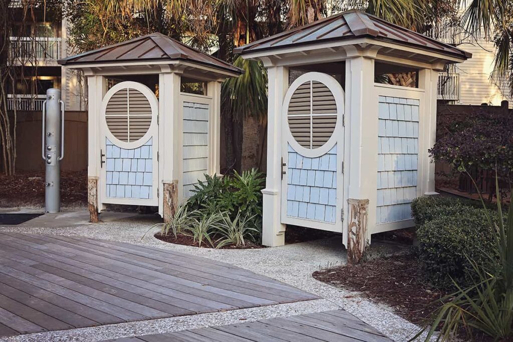 Curved boardwalk with an outdoor shower and two private changing stations, each with a metal roof and doors painted pale blue