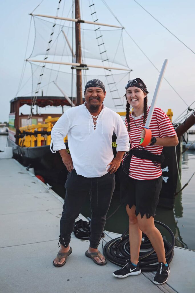 A male and female dressed in pirate gear grin at the camera while standing on a dock with a pirate ship visible in the background