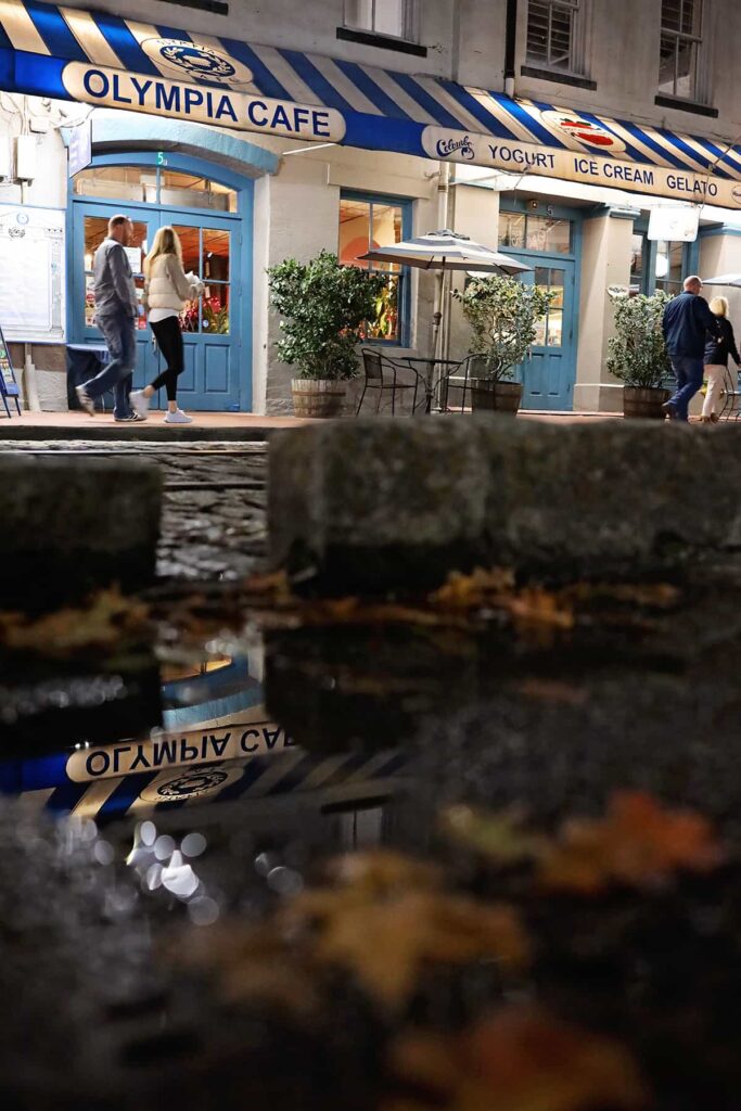Two couples stroll past the blue and white entrance to Olympia Cafe. The sign above the door is reflected in a puddle in the foreground of the image and there are orange and yellow leaves floating in the puddle