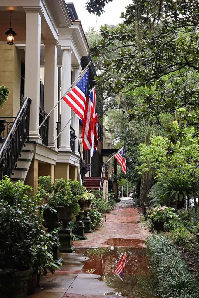View down the sidewalk on Jones Street after a fresh rainfall. Multiple homes have American flags hanging over the sidewalk and lush greenery in planters lining the walkway
