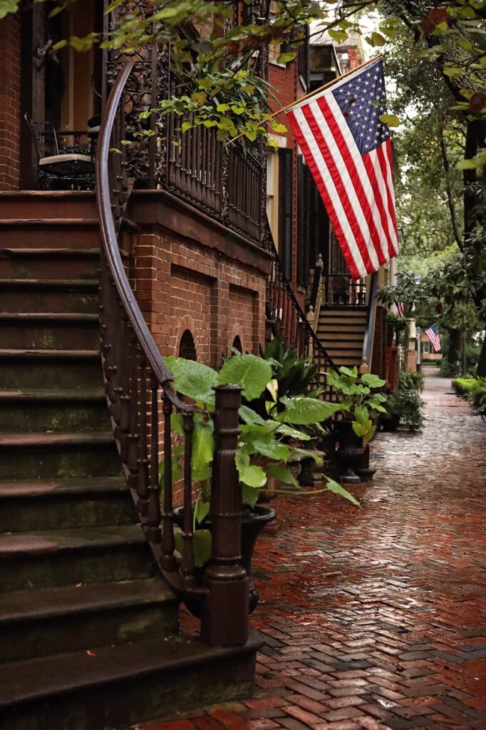 Rainy-day scene in Savannah showing a curved staircase leading to the front door of a stately home. The brick sidewalk in front of the home is laid out in a herringbone pattern and is wet from a recent storm. An American flag hangs from the entryway and lush greenery flanks each side of the stairs