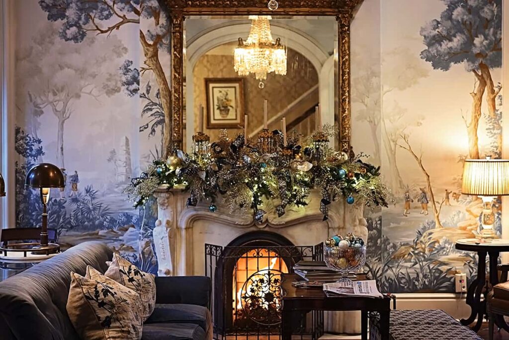 The parlor of Hamilton-Turner Inn shows a cozy seating area in front of a lit fireplace. The mantle is made from carved marble and a gilded mirror rests above it -- reflecting the room's chandelier. A swag of blue and brass ornaments on the mantle matches the designer wall mural
