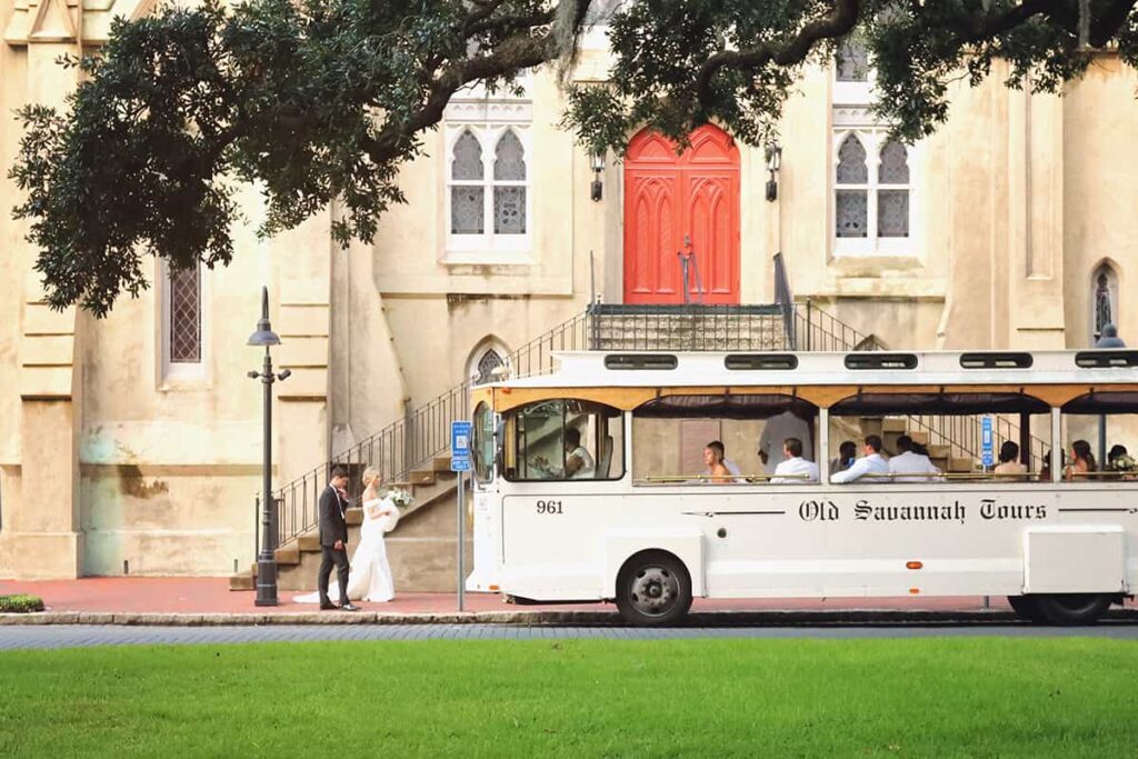 White trolley parked in front of a church with red double doors. A wedding party relaxes in the trolley as an elegant bride and groom walk towards it