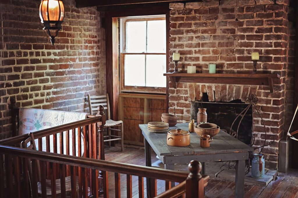 The carriage house of the Sorrel-Weed House is sparsely decorated.The wooden dining table shows a few pottery-style dishes on display, a simple mantle holds four candles, a wooden chair is tucked into one corner, and a quilt is folded and hanging over the stair railing. The walls are all exposed brick