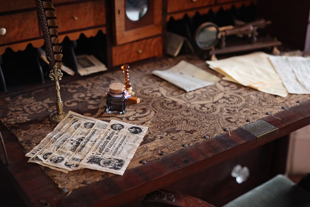 Close-up of a writing desk at the Sorrel-Weed House shows a small display of Confederate bills, scattered papers, and a pen and ink writing device