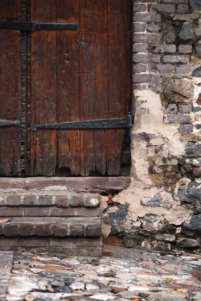 A heavy wooden door on E Factors Walk with hand-forged hinges. The surrounded wall and street in front of the door are made from old ballast stones and the wall is crumbling and damp