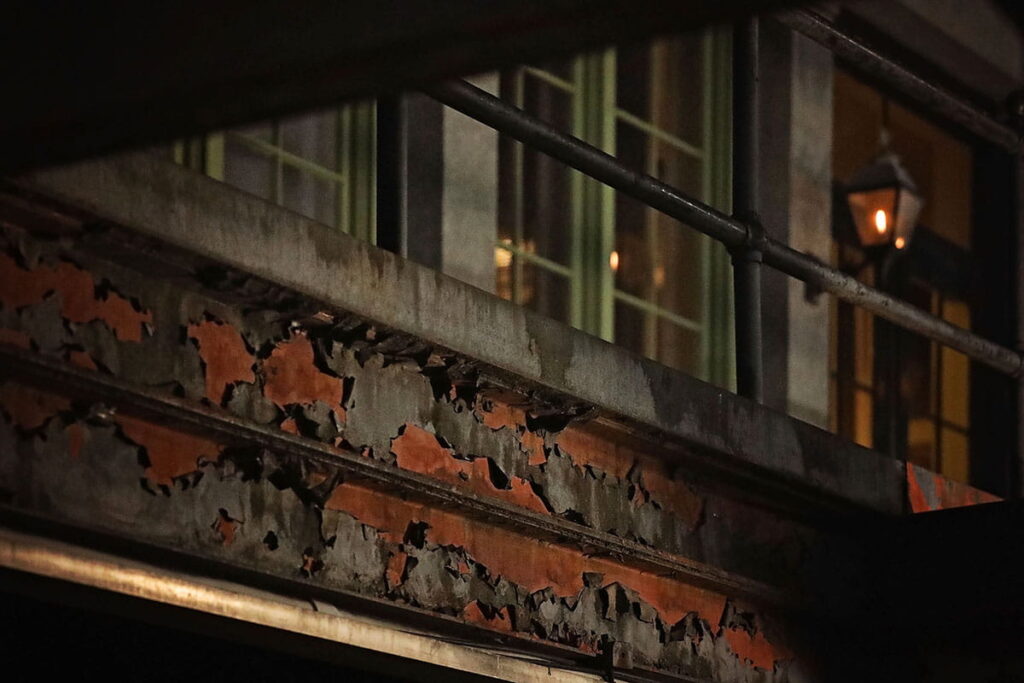 Peeling paint reveals rust on the steel beams supporting the mid level of Factors Walk as a gas lantern glows in the distance